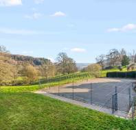 A fenced outdoor tennis court is set on a grassy area with trees and hills in the background under a partly cloudy sky.