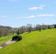 A large, well-kept green lawn with scattered trees, a stone staircase, and a paved path under a clear blue sky with a few clouds.