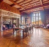 A spacious, upscale library with ornate wooden ceiling, large fireplace, tall bookshelves, wooden floor, and multiple chairs arranged around tables and windows.