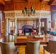 A grand room features a wooden table with chairs, chess sets, an ornate fireplace, wood-paneled ceiling, chandeliers, tall windows, and decorative curtains.