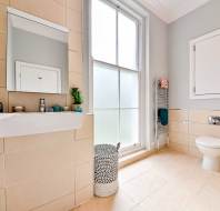 Modern bathroom with beige tiles, a white sink, a wall-mounted mirror, a frosted window, a wicker laundry basket, a towel rack, and a wall-mounted toilet.