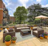 Spacious patio with wicker furniture, a rug, potted flowers, and a dining set with umbrella; surrounded by a metal railing and overlooking trees and brick buildings.