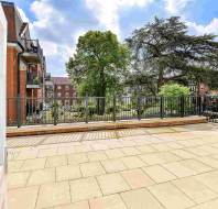 Spacious paved terrace with a metal railing overlooks a landscaped garden with trees and residential brick buildings in the background.