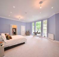 Spacious bedroom with light purple walls, large windows, a double bed, a small desk near the windows, and an open closet with hanging clothes.