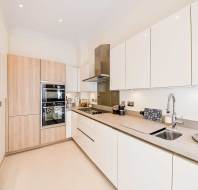 Modern kitchen with white cabinets, built-in oven, electric cooktop, stainless steel hood, beige countertops, and various utensils on the counter.