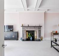 A spacious living room with a marble fireplace, a black TV cabinet, a glass coffee table, and light pink walls. The ceiling features decorative white beams.