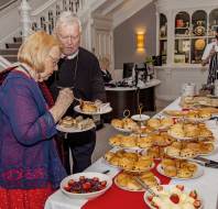 Elderly man and woman serve themselves pastries and desserts from a buffet table in a well-decorated room, with staff and more food visible in the background.