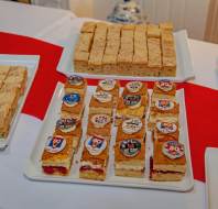 Three trays of rectangular cakes and one tray of layered cakes topped with circular edible VE Day decorations are displayed on a table with a red and white tablecloth.