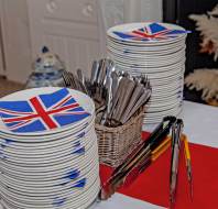Stacks of plates with Union Jack napkins, a basket of cutlery, and serving tongs are arranged on a table with a red and white tablecloth.