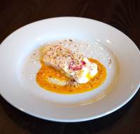 A rectangular slice of dessert topped with crushed nuts sits on a white plate, surrounded by a yellow-orange sauce on a dark wooden table.