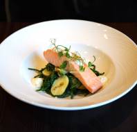A plated dish with a piece of cooked salmon on a bed of green vegetables, garnished with microgreens and accompanied by small dollops of sauce.