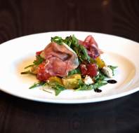 A white plate with prosciutto, arugula, yellow and red tomatoes, crumbled cheese, and balsamic glaze on a dark wooden table.