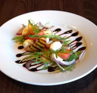 A white plate with grilled vegetables, cherry tomatoes, mozzarella, red onion, arugula, and balsamic glaze on a dark wooden table.