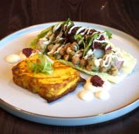 A plated dish with a chickpea and vegetable salad on lettuce, garnished with sauce and herbs, served alongside a golden rectangular baked item and small sauce dollops.