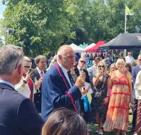 A man in a blue suit speaks into a microphone outdoors, surrounded by a crowd of people at an event with tents and trees in the background.
