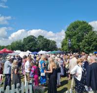A large group of people in summer attire gather outdoors near white tents and a picket fence, with trees and a blue sky in the background.