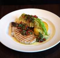 A white plate with a grilled fish fillet, grilled lettuce, two boiled potatoes, and a green herb sauce garnished with diced red peppers.