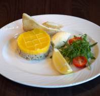 A plated dish with a round yellow-topped pâté, two toast slices, a scoop of butter, cherry tomatoes, arugula, and a lemon wedge on a white plate.