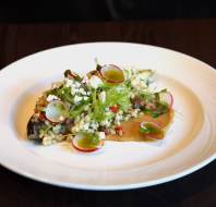 A plated dish featuring grains, sliced radish, greens, and cheese on a toasted flatbread, presented on a white plate against a dark table.
