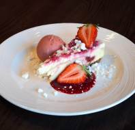 A slice of cheesecake with strawberry sauce, fresh strawberries, crumbled white topping, and a scoop of pink ice cream on a white plate.