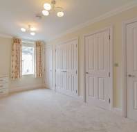 A bright, empty room with beige carpet, pale walls, three closed white doors, a window with patterned curtains, and built-in white drawers under the window.