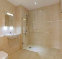 Accessible bathroom with beige tiles, wall-mounted sink, open roll-in shower, grab bar, handheld showerhead, mirror, and heated towel rail.