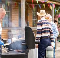 A man in an apron grills food outdoors while two older men stand nearby. Colorful bunting decorates a wooden building in the background.