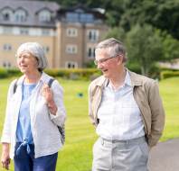 Two older adults, a woman and a man, walk outdoors on a grassy area near a large building on a sunny day.