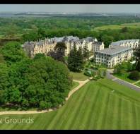 Aerial view of a large historic building surrounded by green lawns, trees, and walking paths, with countryside visible in the distance. Text reads "The House & Grounds.