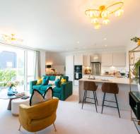 Modern open-plan living room and kitchen with large windows, green sofa, yellow armchair, breakfast bar with stools, and bright, neutral decor.