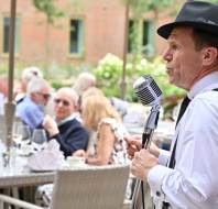 A man in a white shirt, black tie, and hat sings into a vintage microphone at an outdoor event with people seated at tables in the background.
