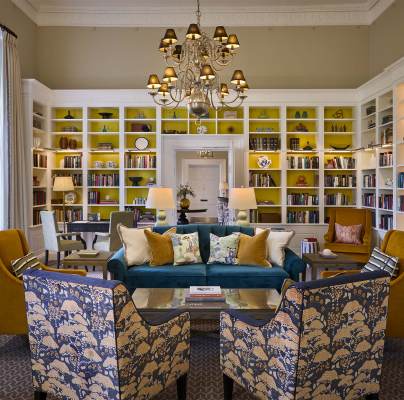 Elegant library room with a chandelier, large yellow bookshelves, a green sofa, patterned armchairs, and a central coffee table, creating a cozy reading area.