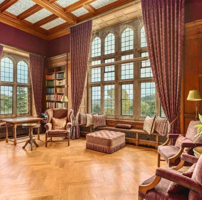 A spacious, elegant library with tall windows, purple drapes, wooden floors, bookshelves, and comfortable seating.