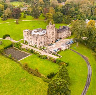 Aerial view of a large stone castle surrounded by green lawns and trees, with a driveway and parked cars in front. A tennis court is visible on the left.