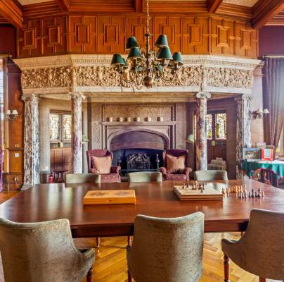 A grand room features a wooden table with chairs, chess sets, an ornate fireplace, wood-paneled ceiling, chandeliers, tall windows, and decorative curtains.