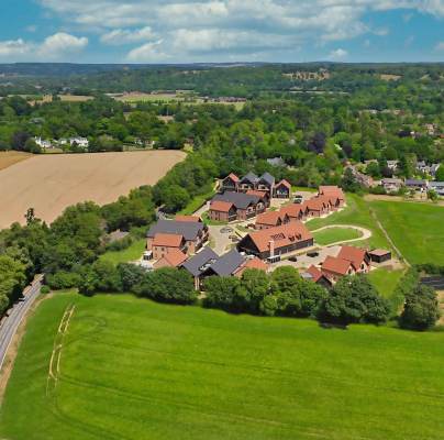 Aerial view of a rural area with fields, trees, and a cluster of buildings in the center, surrounded by greenery and distant houses under a partly cloudy sky.