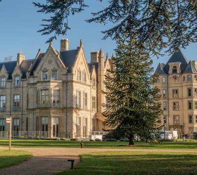 A large historic building with ornate architecture is surrounded by a grassy area, trees, and a clear blue sky.