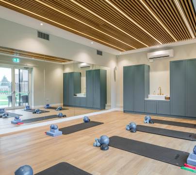 Modern fitness studio with wooden flooring, mirrored wall, ceiling slats, exercise mats, and props arranged for a class. Cabinets and a sink are visible at the back of the room.