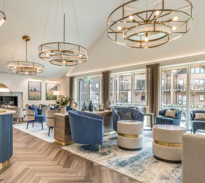 A modern lounge with blue and beige armchairs, round tables, gold light fixtures, large windows, and wood-patterned flooring.