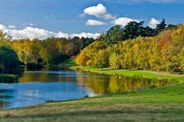 A calm lake bordered by grassy fields and trees with green and yellow foliage under a blue sky with scattered clouds.