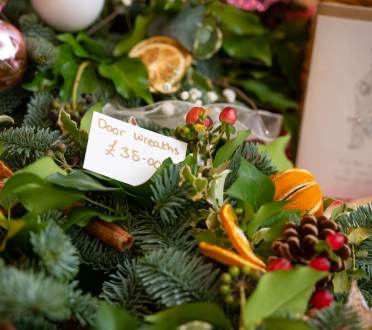 A close-up of festive door wreaths decorated with greenery, berries, dried orange slices, and pinecones, with a handwritten sign reading "Door Wreaths £35.00".