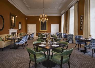 The Romney at Mote House Elegant dining room with green and blue chairs, set tables, chandeliers, and paintings on warm brown walls. Natural light streams through large windows with beige curtains.