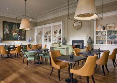 The Romney at Mote House A modern cafe with brown and green chairs, wooden tables, large pendant lights, and wall art. Shelves display decorative vases.