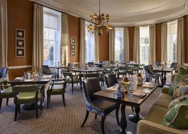 Elegant dining room with neatly arranged tables and chairs. Large windows with curtains and a chandelier overhead provide soft lighting. Neutral tones with green and brown accents throughout.
