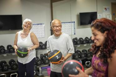 Three people are joyfully exercising with medicine balls in a gym, surrounded by dumbbells and a mirror.