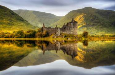 Ancient stone castle ruins stand by a calm lake, reflecting in the water, with green hills and cloudy sky in the background.