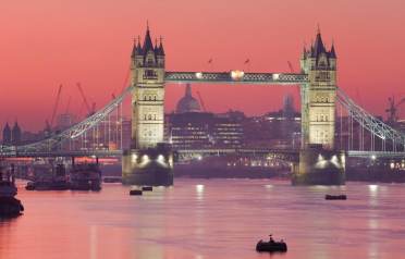 Tower Bridge in London is illuminated at dusk, with calm water in the foreground and the city skyline visible under a pink-orange sky.