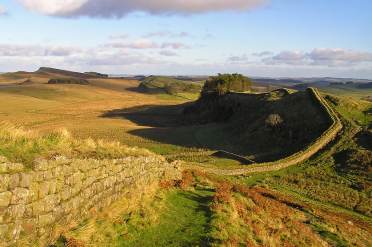 A stone wall winds across a grassy, hilly landscape under a blue sky with scattered clouds. Shadows from the hills and trees are visible on the ground.