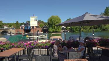 Two people sit at an outdoor riverside café table under an umbrella, with boats, flowers, and buildings visible across the water on a sunny day.
