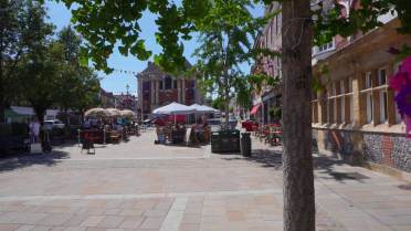 Outdoor dining area on a sunlit pedestrian street with people sitting at tables under umbrellas, surrounded by trees and buildings.